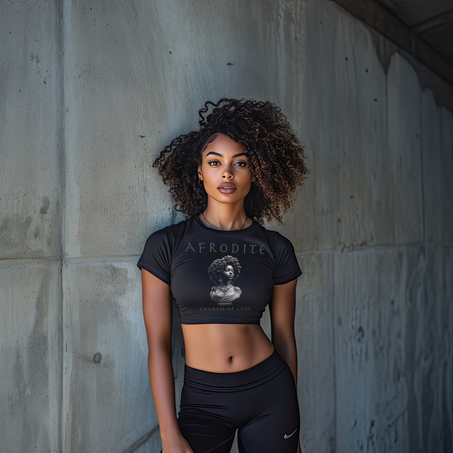 "Black woman wearing Afrodite crop top with goddess graphic, standing confidently against a concrete wall."